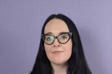 Eine weiblich gelesene Person vor lilanem Hintergrund, mit schwarzen langen Haaren und schwarzer Brille in einem weißen T-shirt.