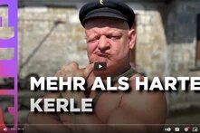 Eine männlich-gelesene Person mit Glatze und Hut macht eine Grimasse in die Kamera, oberkörperfrei flext die Person den Arm. In weiß steht davor "Mehr als harte Kerle".