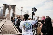 Mehrere Menschen von hinten auf der Brooklyn Bridge. Eine als schwarz gelesene Person hält ein Megafon in die Luft.