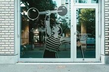 Aufkleber einer als männlich gelesenen Person in gestreiftem Top, die eine Hantel in die Luft streckt, aufgeklebt auf einer Glastuer