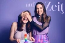 Die beiden Postcast-Hosts stehen vor lila Hintergrund, die rechte Person hält der linken die Augen zu,  beide lachen. Darüber in geschwungener Schrift der Titel des Podcasts