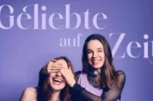 Die beiden Postcast-Hosts stehen vor lila Hintergrund, die rechte Person hält der linken die Augen zu,  beide lachen. Darüber in geschwungener Schrift der Titel des Podcasts