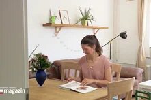 Screenshot des TV-Beitrags "Jede dritte Frau erlebt Gewalt während der Geburt". Eine als Frau gelesene Person in einem rosafarbenen Oberteil sitzt an einem Holztisch und blättert in einem Buch. Im Hintergrund stehen eine blaue Vase mit Blumen, ein Regal mit Bildern und eine gemütliche Wohnzimmereinrichtung.