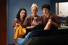 Drei Personen in braunen Arbeitskleidern stehen in einem medizinischen Raum. Eine hält ein Neugeborenes, eine weitere ein gelbes Handtuch. Thema: Geburt und medizinische Betreuung.