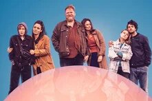 Sechs Personen stehen eng beieinander vor einem pinkfarbenen Hintergrund. Sie wirken verbunden und selbstbewusst.