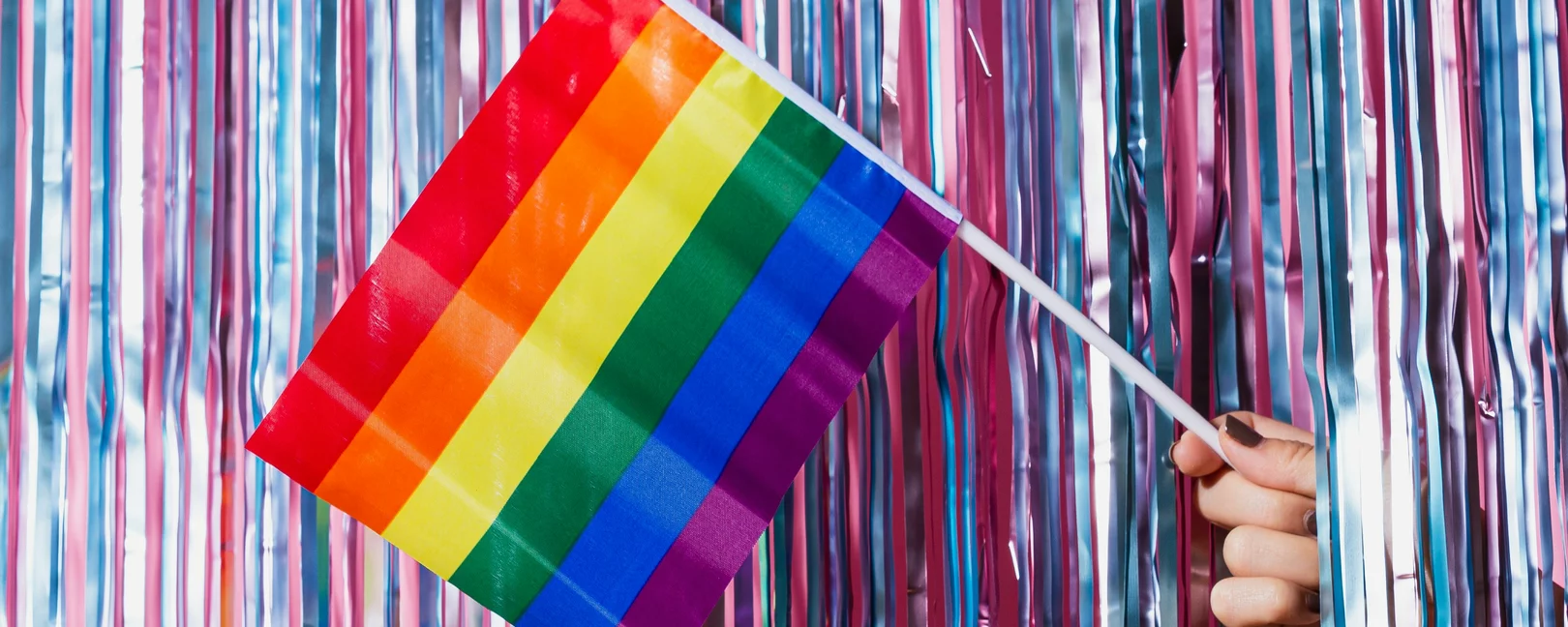 Vor einem Lamettavorhang wird eine Prideflagge rausgehalten.