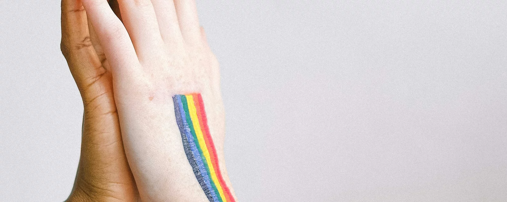Zwei Personen stehen nah beieinander und berühren ihre Hände. Auf dem Arm ist eine Regenbogenflagge.