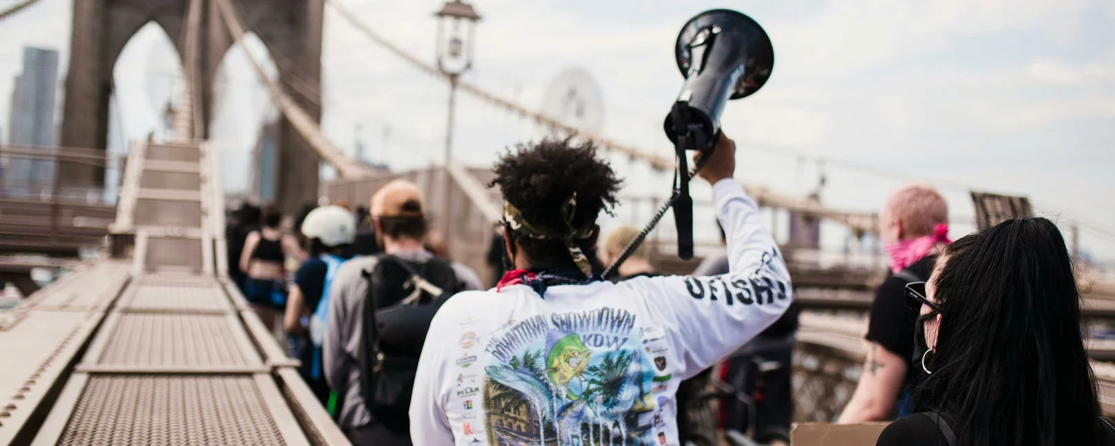Mehrere Menschen von hinten auf der Brooklyn Bridge. Eine als schwarz gelesene Person hält ein Megafon in die Luft.
