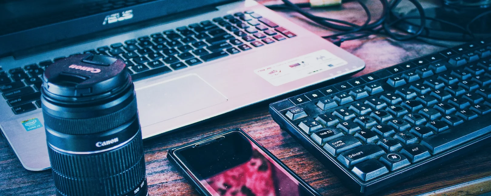 Laptop, Tastatur, Kamera, Handy liegen auf einem Holztisch, rosa Licht scheint hervor