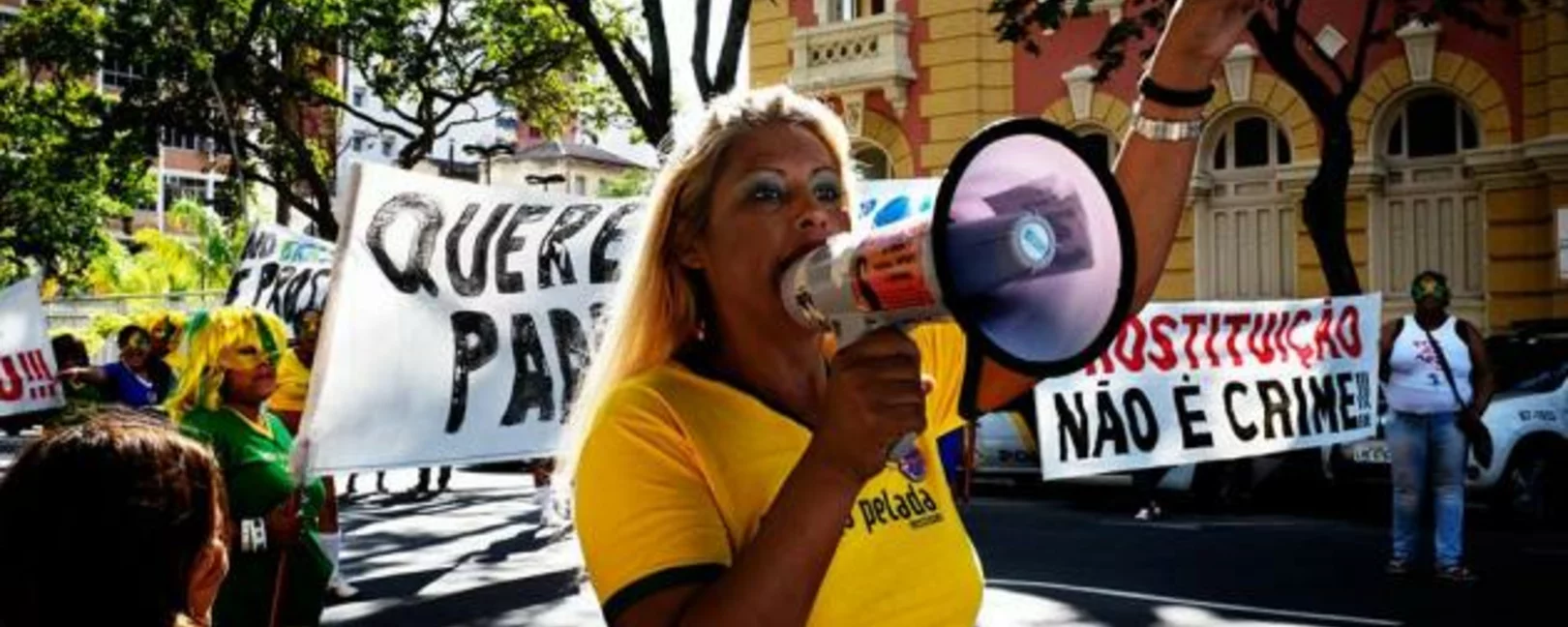 eine Person mit einem gelben T-Shirt und langen Haaren spricht auf einer Demo in ein Megafon